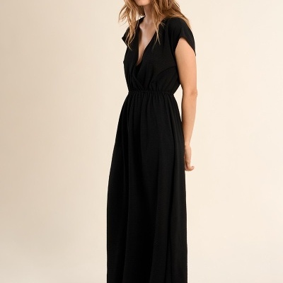 Mulher com vestido longo preto de mangas curtas e decote em V, fundo bege.