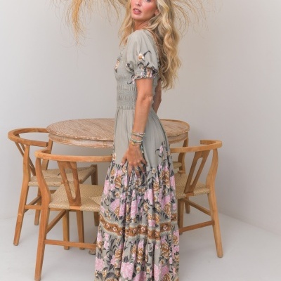 Vestido comprido cinzento com flores rosa e castanhas usado por mulher ao lado de mesa e cadeiras de madeira