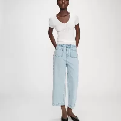 Mulher vestindo camisola branca de malha, jeans largos azul claro, meias castanhas transparentes e sapatos pretos de salto baixo