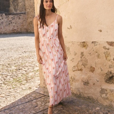 Vestido longo de alças finas com padrão floral e riscas, com sandálias castanhas, junto a parede de pedra.