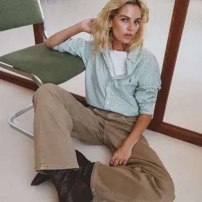 Mulher sentada no chão com camisa às riscas verde claro e branco, calças bege largas e botas castanhas, ao lado de cadeira verde com estrutura metálica.
