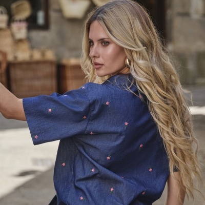 Camisa azul com pequenas flores cor-de-rosa bordadas, usada por mulher loira com brincos dourados.