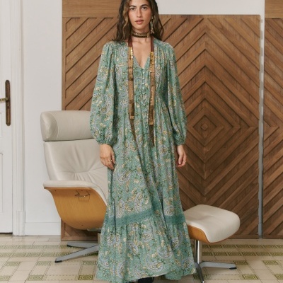 Mulher com vestido comprido verde com padrão cachemira em ambiente interior com cadeira e painel de madeira.