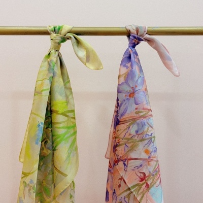 Dois lenços florais em tons verdes/amarelos e roxos/azuis pendurados numa barra amarela.