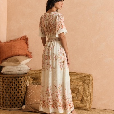 Vestido comprido branco com flores cor-de-rosa em modelo feminino de costas, com sapatos e decoração rústica ao fundo.