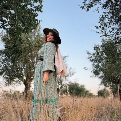 Mulher com vestido longo verde e chapéu verde em campo aberto com relva seca e árvores ao fundo.