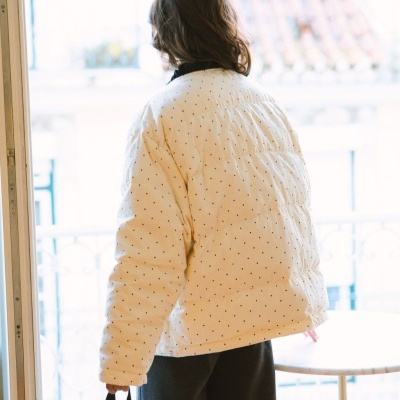 Casaco creme acolchoado com pontos pretos usado por pessoa junto a janela com bolsa peluda