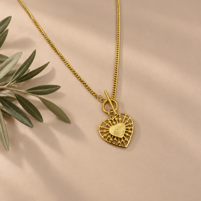 Colar dourado com pendente em forma de coração e corrente sobre fundo bege com folhas verdes ao lado