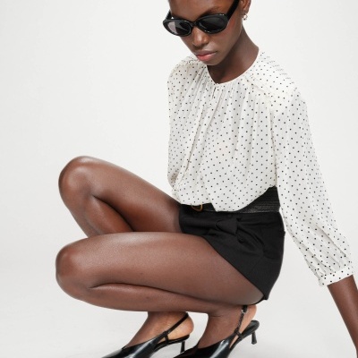 Mulher com blusa branca de pontos pretos, calções pretos e sapatos de salto baixo pretos, usando óculos de sol.