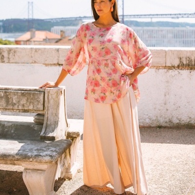 Mulher veste blusa floral rosa e calças largas creme junto a banco de pedra num espaço exterior com ponte ao fundo.