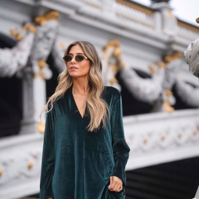 Mulher com vestido de veludo verde escuro e óculos de sol, segurando mala castanha, com ponte branca decorada ao fundo