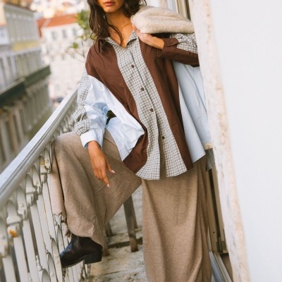 Mulher com camisa bicolor azul claro e castanha, saia comprida castanha clara, botas pretas e casaco de malha bege numa varanda