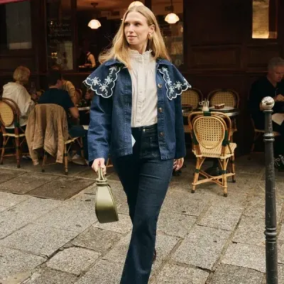 Mulher com casaco de ganga azul com bordados brancos, camisa branca, jeans escuros, botas castanhas e mala verde num passeio à frente de esplanada.