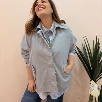 Mulher com casaco azul claro e camisa azul e branca, calças jeans, ao lado de planta decorativa com cartão