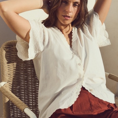 Blusa branca com renda nas mangas e calças castanhas, modelo sentado numa cadeira de vime