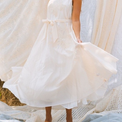 Vestido branco comprido com bordados florais e cinto, sandálias castanhas, com tecidos brancos ao fundo, texto maïté