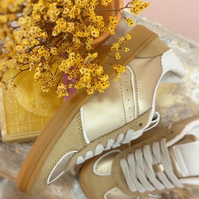 Ténis dourados e beges com atacadores brancos e sola castanha sobre mesa com flores secas amarelas