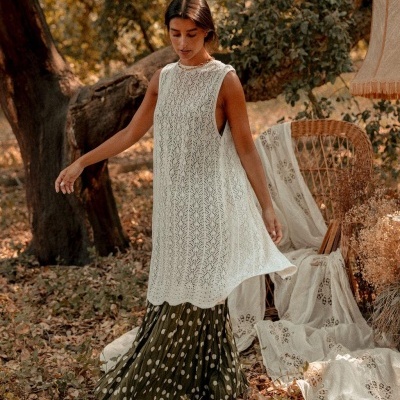 Mulher com vestido branco com padrão de renda e saia verde com bolinhas brancas em ambiente exterior com folhas secas e cadeira de vime