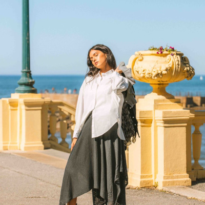 Mulher vestida com camisa branca, saia cinza assimétrica e botas pretas ao ar livre junto ao mar