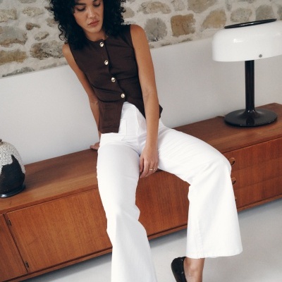 Mulher sentada com top castanho e calças brancas numa cómoda de madeira