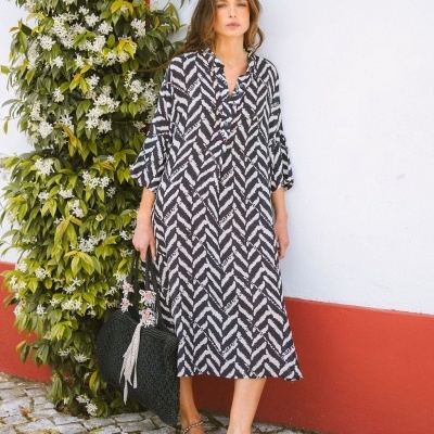 Mulher com vestido midi preto e branco, segurando mala preta, junto a parede branca e vermelha com plantas