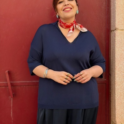 Mulher vestida com camisola azul escura, calças pretas, lenço vermelho, joias douradas e brincos verdes, sorrindo diante de porta vermelha.