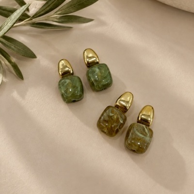 Dois pares de brincos dourados com pendentes quadrados em resina marmoreada verde e amarela sobre fundo bege e folhas verdes ao lado.