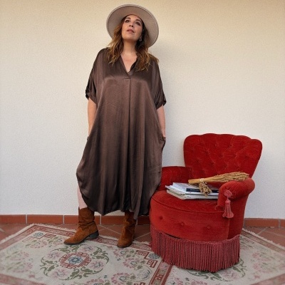 Mulher de vestido castanho fluído, chapéu branco, botas castanhas, ao lado de poltrona vermelha e livros.