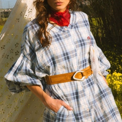 Mulher com vestido xadrez azul e branco, cinto castanho e lenço vermelho ao pescoço em campo florido