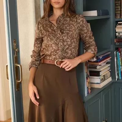 Mulher com camisa floral castanha e saia castanha junto a estante azul esverdeada