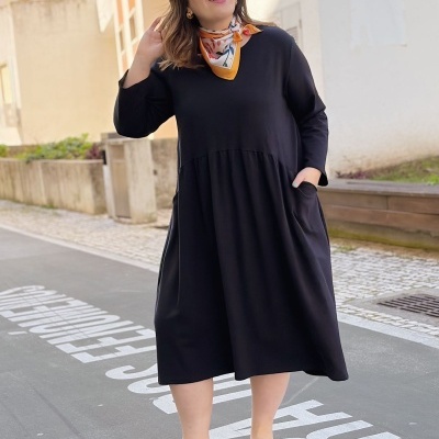 Mulher com vestido midi preto de manga comprida e lenço colorido no pescoço em ambiente urbano.