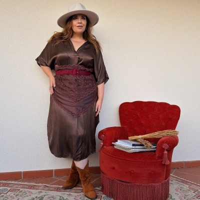 Mulher com vestido castanho acetinado e chapéu bege junto a poltrona vermelha com revistas.