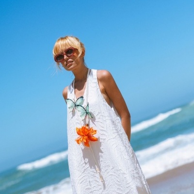 Mulher com vestido branco longo, colar com flor laranja e borboletas, óculos escuros, praia ao fundo.