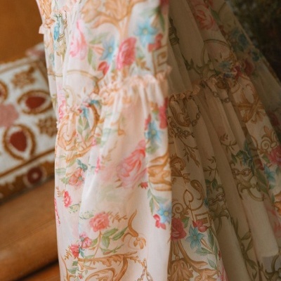 Vestido floral com padrão em tons pastel e detalhes dourados junto a sofá castanho.
