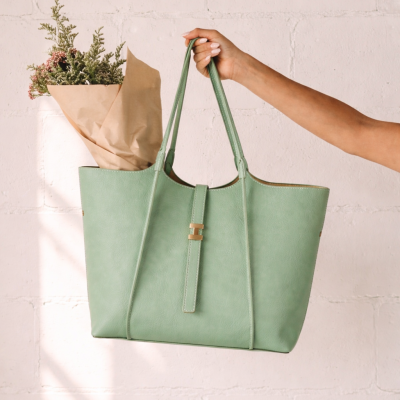 Saco de mão verde claro com flores dentro, segurado por mão feminina contra parede branca.