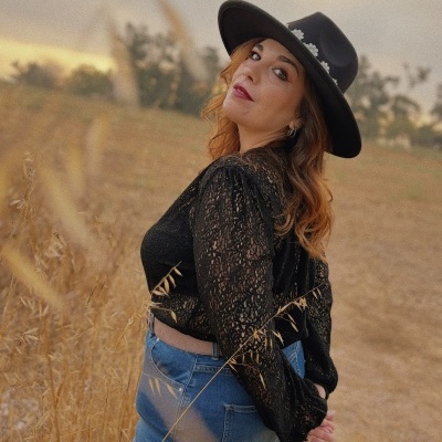 Mulher com chapéu preto decorado com flores brancas, blusa preta de renda e calças de ganga em campo seco ao entardecer