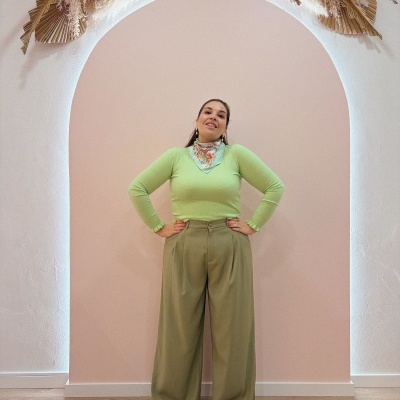 Mulher vestida com roupa verde e calças largas em fundo de parede rosa com arco decorado.