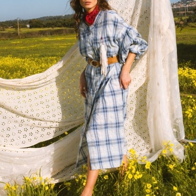 Mulher com vestido xadrez azul e branco e lenço vermelho num campo de flores amarelas.