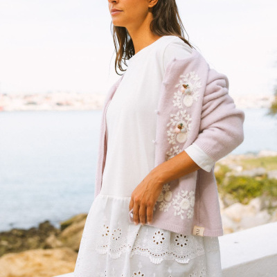 Mulher veste macacão branco bordado e casaco rosa claro com bordados florais junto ao mar.