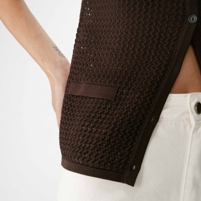 Colete castanho escuro em malha com padrão ondulado e bolso lateral perto de calça clara com botão dourado