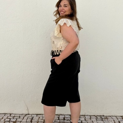 Mulher de perfil com blusa branca, saia ou calças pretas, cinto, sandálias pretas, em fundo branco e chão de calçada