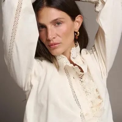 Blusa creme feminina com renda e botões, usada por modelo com cabelo castanho e brincos dourados.