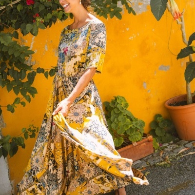Mulher com vestido longo floral amarelo ao ar livre com parede amarela e plantas