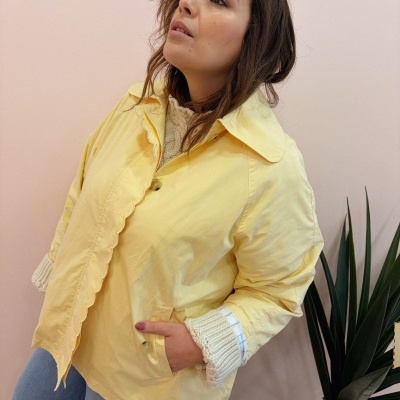 Mulher com casaco amarelo e camisola de malha creme em fundo bege com planta