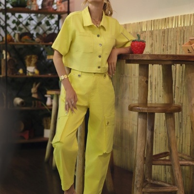 Conjunto feminino amarelo com camisa de manga curta e calças cargo, sandálias castanhas, ambiente interior com móveis rústicos e plantas.