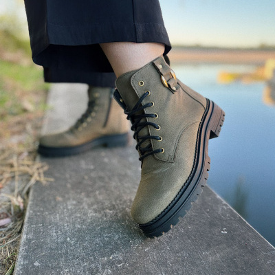 Bota verde-oliva de camurça com sola grossa e fivela dourada, usada com calças pretas perto de água.