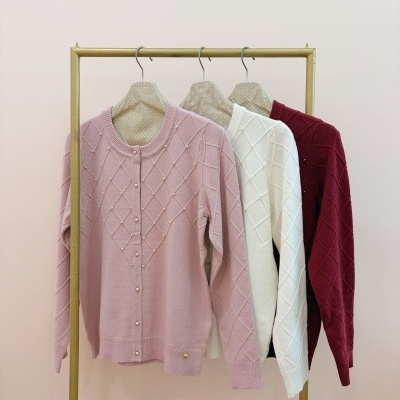 Três cardigans rosa, branco e vinho pendurados em cabides num suporte metálico dourado