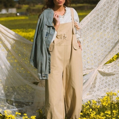 Mulher de macacão bege com bordado floral, camisa branca e casaco de ganga em campo florido amarelo.