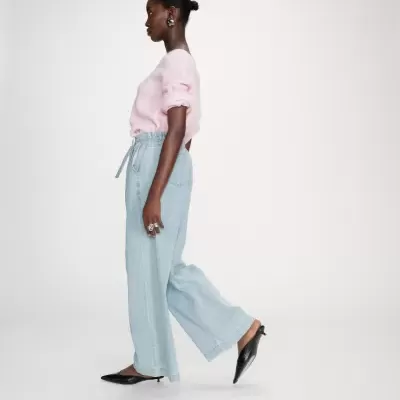 Mulher de perfil com camisola rosa, calças largas em ganga azul claro e sapatos pretos.