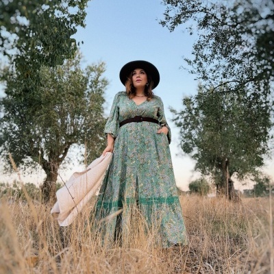 Mulher com vestido verde comprido e chapéu preto segurando casaco bege ao ar livre entre árvores